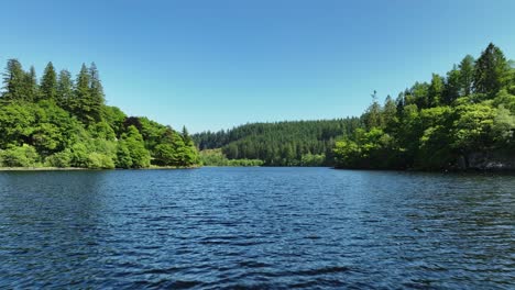 Aerial-drone-footage-flying-over-the-surface-of-still-water-of-Loch-Ard-in-the-Troasachs-National-Park-Scotland-towards-a-native-scots-pine-forest-with-clear-blue-sky-on-a-sunny-day