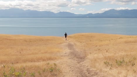 Mann-Rennt-Die-Goldene-Helliwell-Wiese-Hinunter-In-Richtung-Meer-Mit-Den-Felsigen-Bergen-Im-Hintergrund-Auf-Hornby-Island-In-British-Columbia,-Kanada