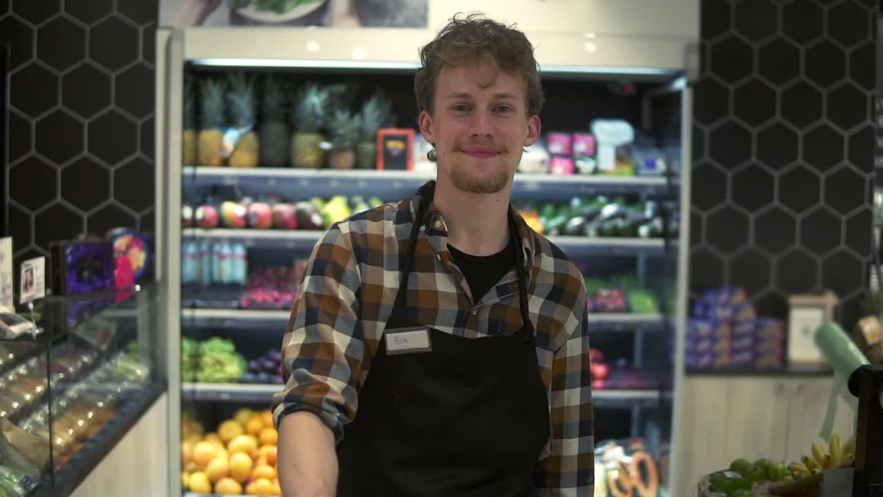 retrato del joven guapo trabajador de la tienda caucásica en el delantal frente a la cámara y sonriendo alegremente mientras empuja el carrito en el supermercado de grosería