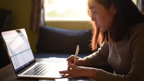 focused individual engaged in detailed work on laptop, taking notes with pen at home during bright afternoon light