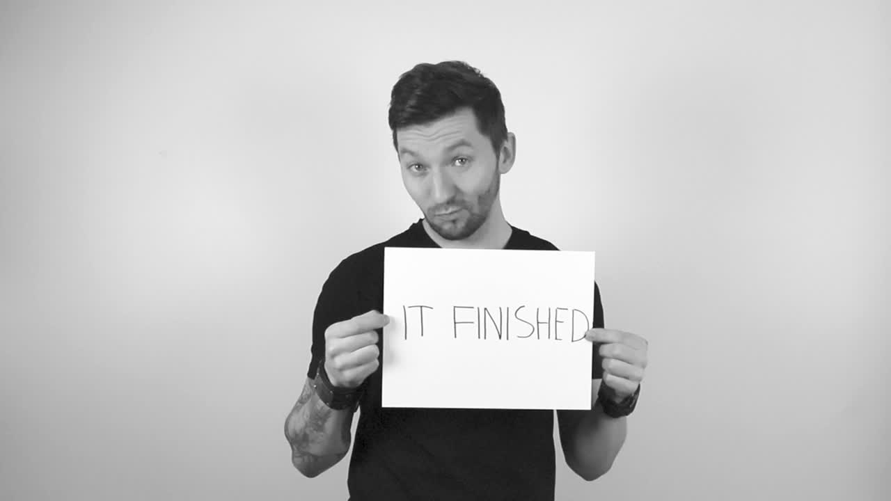 Grayscale footage of a tattooed man with short hair and trimmed beard raising up or lifting up a white paper sign in his hands that says It Finished in capital letters while being happy and confident.