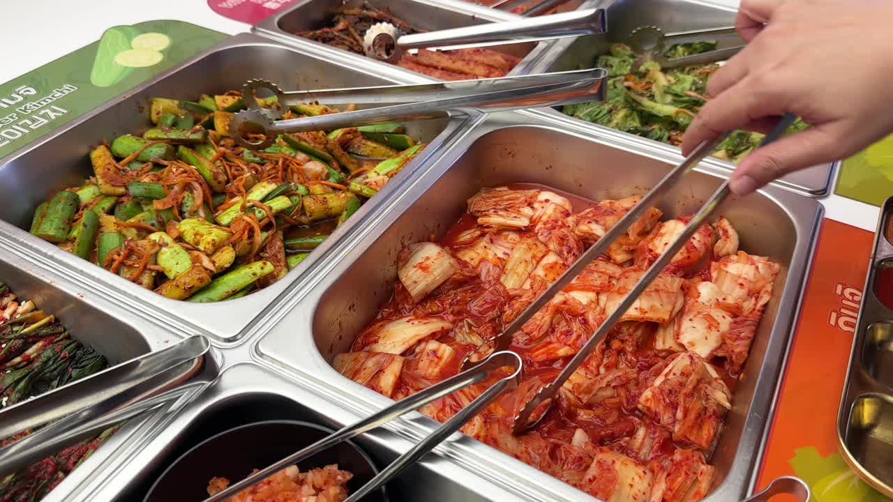 Hand uses tongs to pick kimchi from buffet with assorted Korean side dishes, bright lighting