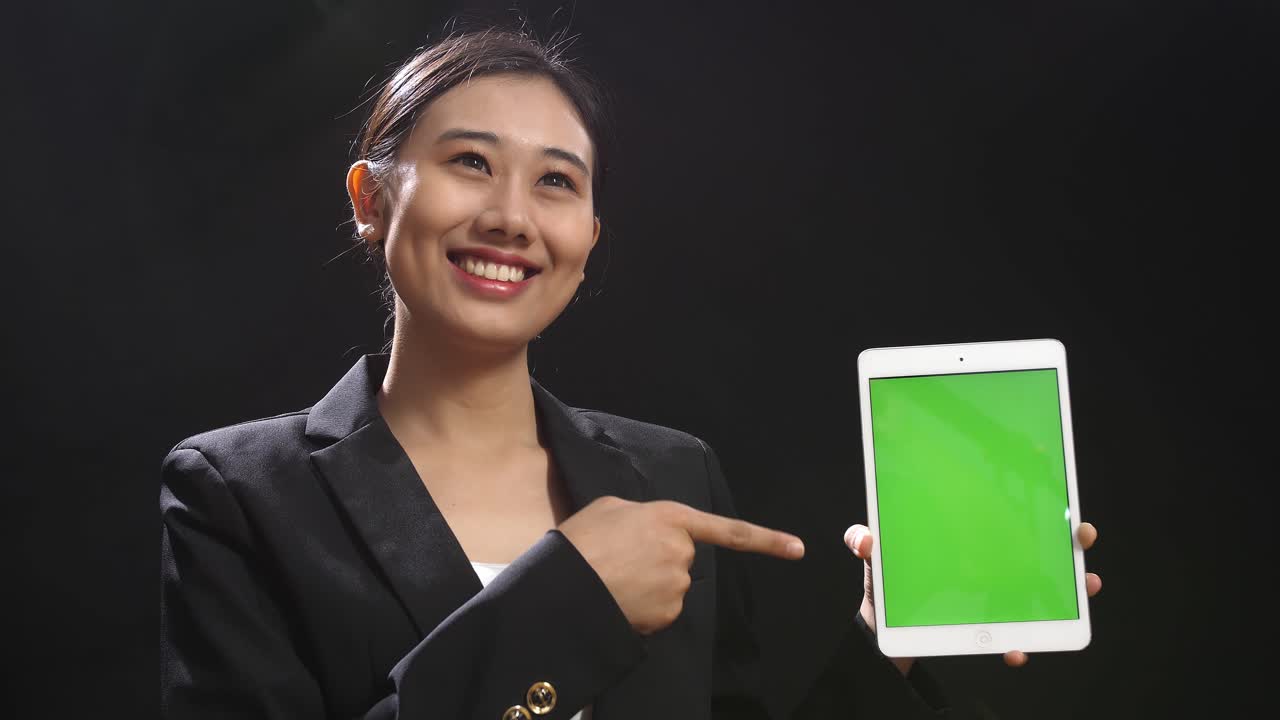 블랙 스크린 스튜디오에서 녹색 스크린 태블릿을 들고 가리키는 비즈니스 슈트를 입은 아시아 스피커 여성의 클로즈업