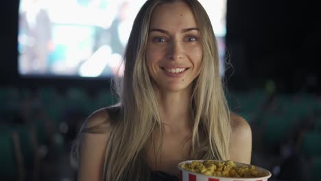 Blonde-woman-standing-with-bucket-of-popcorn-at-the-cinema,-smiling-to-the-camera