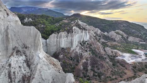 jagged white cliffs contrasted by a lush green pine forest, set against a backdrop of dark, dramatic storm clouds in western greece. dolly forward