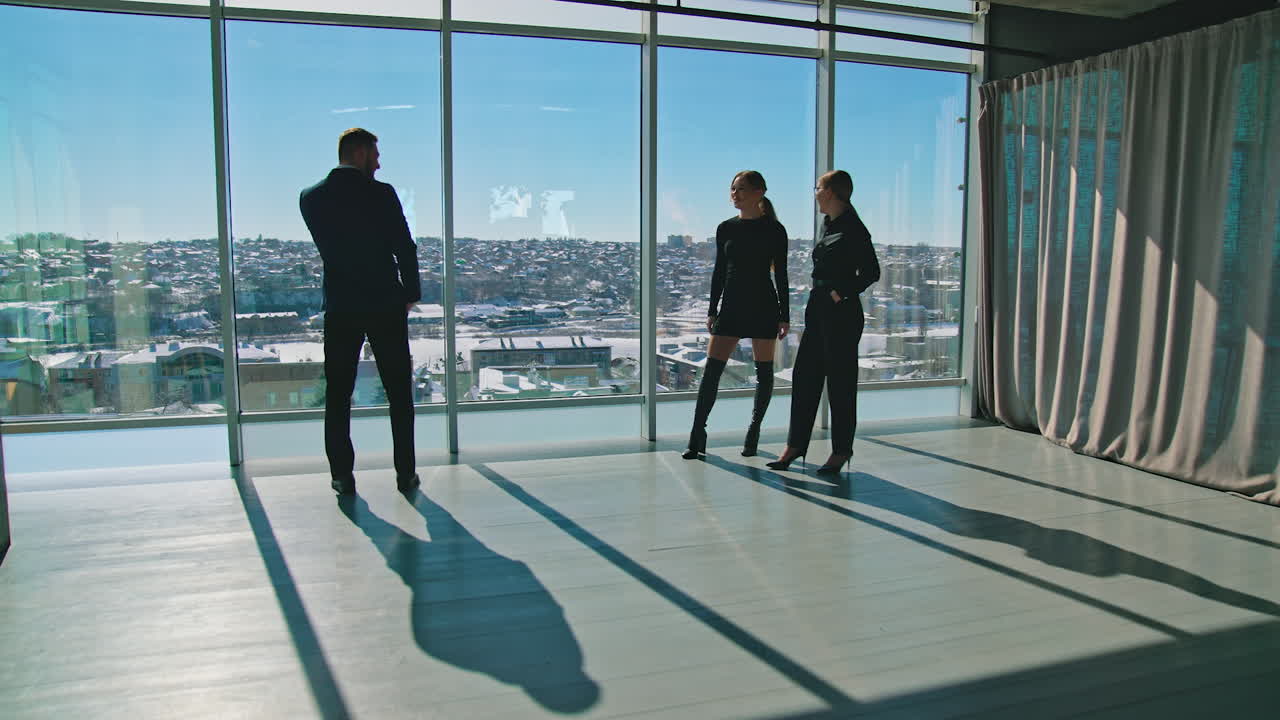 Male entrepreneur in suit with a phone and women indoors. Beautiful young female partners waving hello to a businessman standing near the window.
