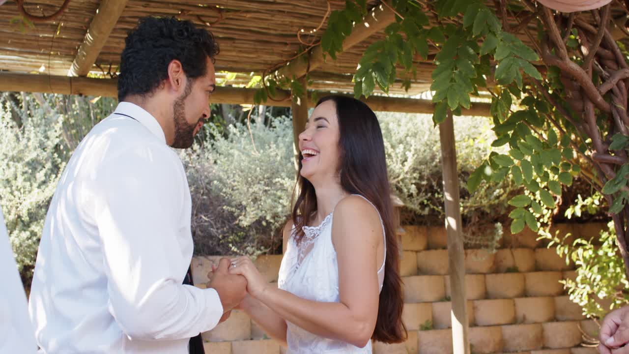 Bride and groom laughing, holding hands under pergola, officiant inviting vows, groom placing ring