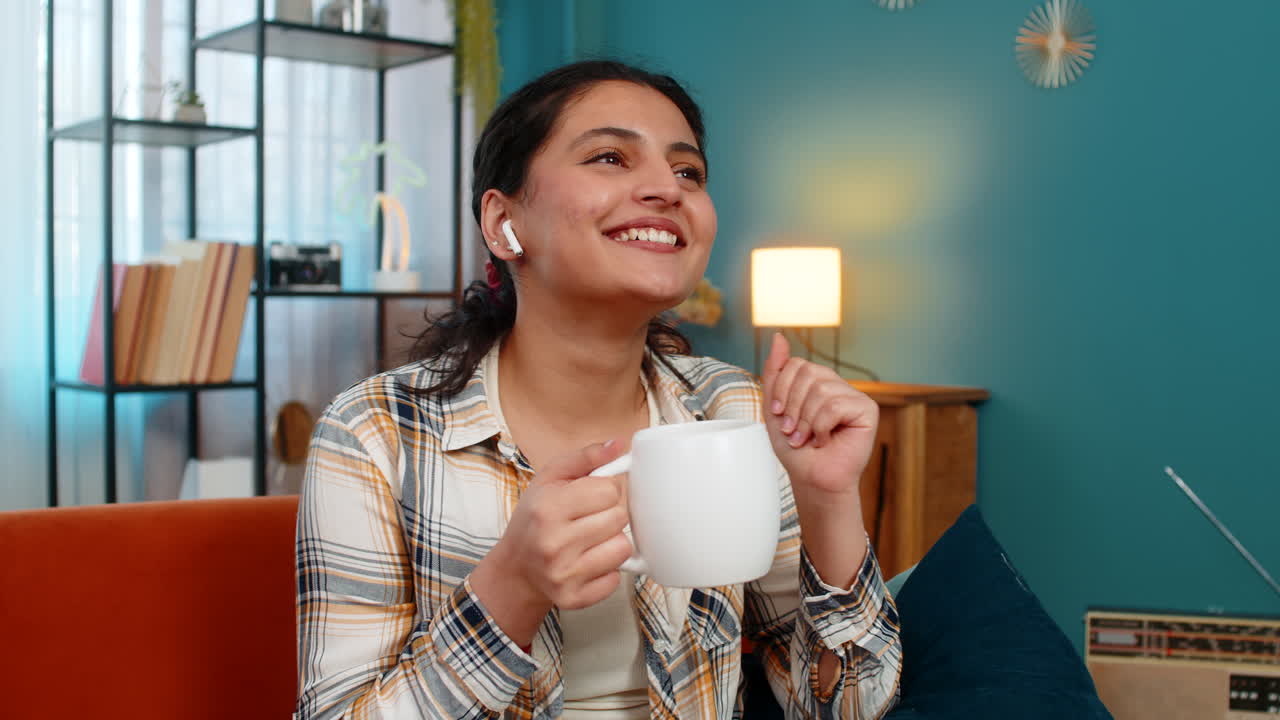 Mujer india con auriculares escuchando música calmante y relajante sosteniendo una taza de café sin preocupaciones en casa