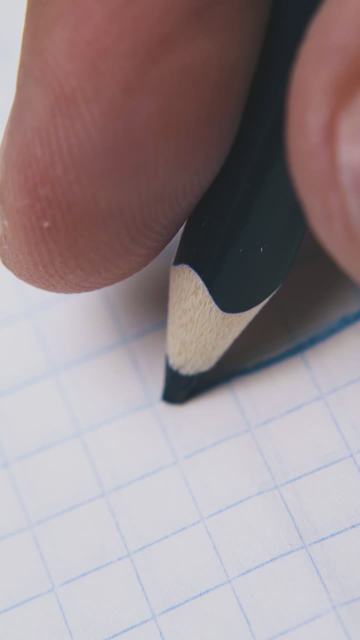 young man draws straight line using dark blue pencil on checkered paper sheet extreme close view