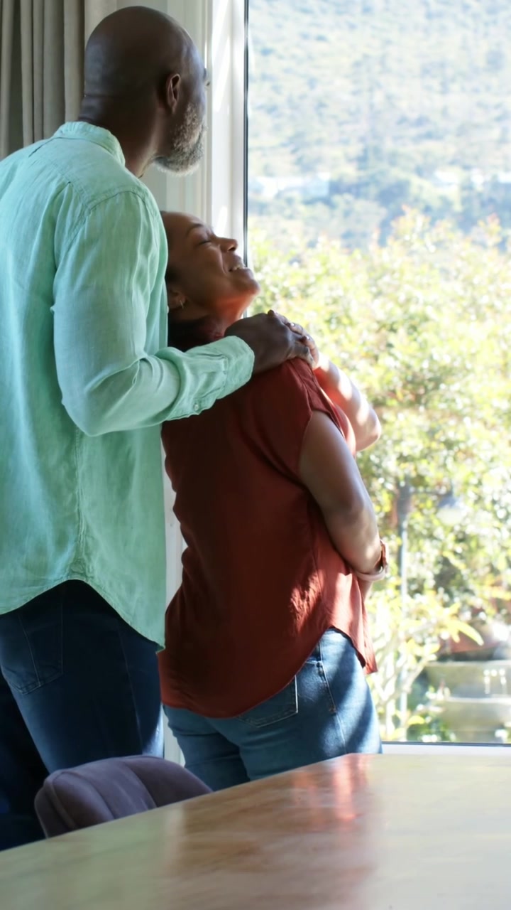 Video verticale: Uomo che entra da sinistra e tocca una donna con una camicetta color ruggine, appoggiata alla finestra per comodità.