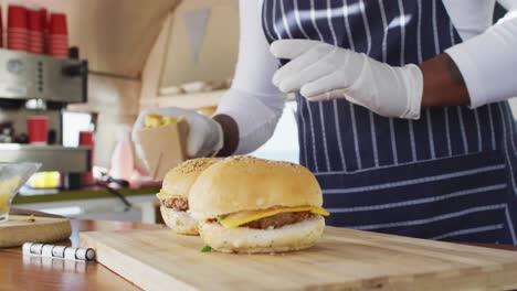 mid section of african american man wearing apron putting burgers and fries over wooden tray