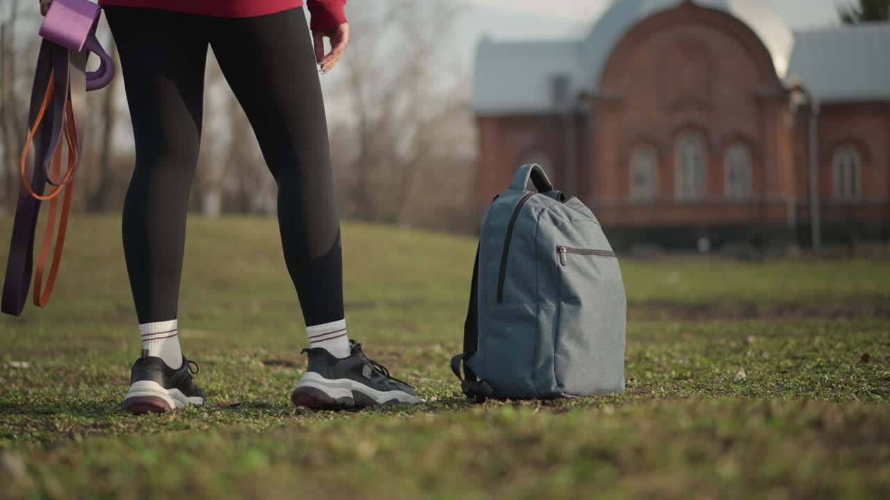 Lady Strolling With Leash Near Structure, Female Individual With Bag Moving Through Grassy Area, Woman Carrying Backpack Walking Past Brick Wall In Grassy Setting During Daily Routine