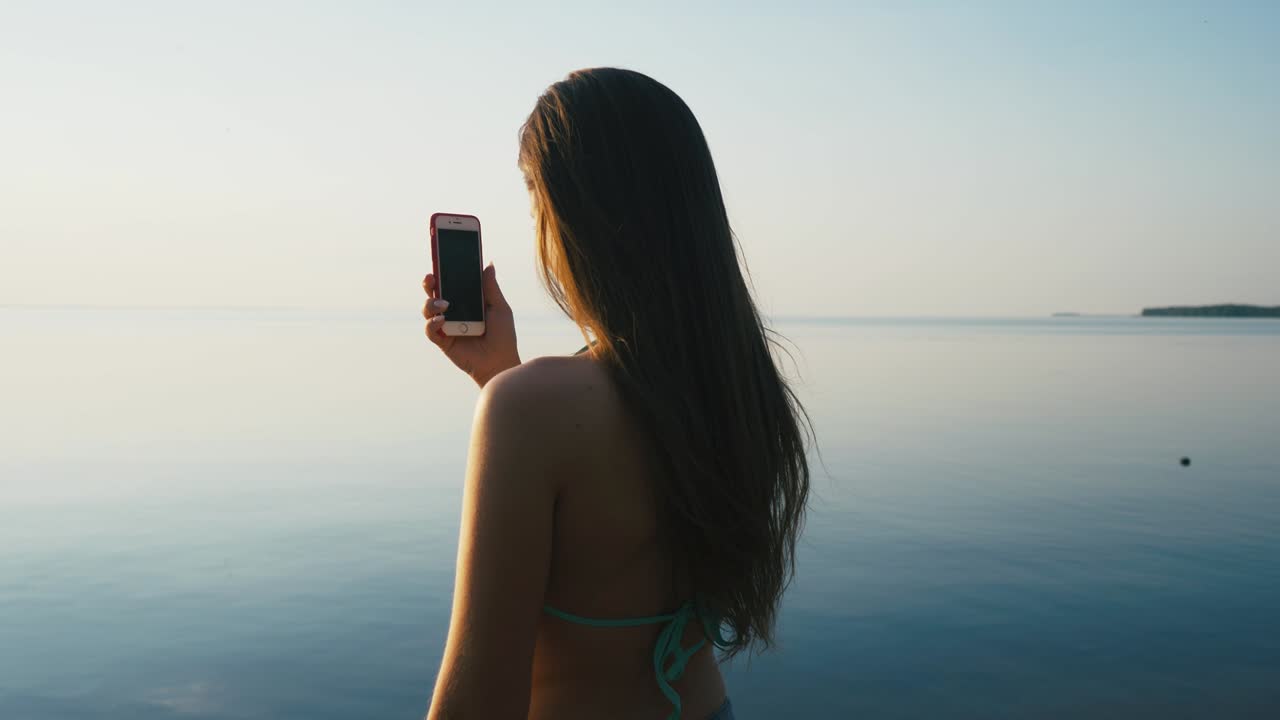 아름다운 젊은 여성이 휴가 중 해변 근처의 해가 지는 동안 스마트폰으로 사진을 찍고 있습니다.