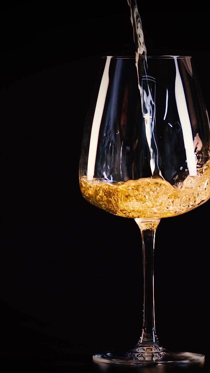 Un flusso di vino bianco dorato riempie un bicchiere di cristallo su uno sfondo scuro.