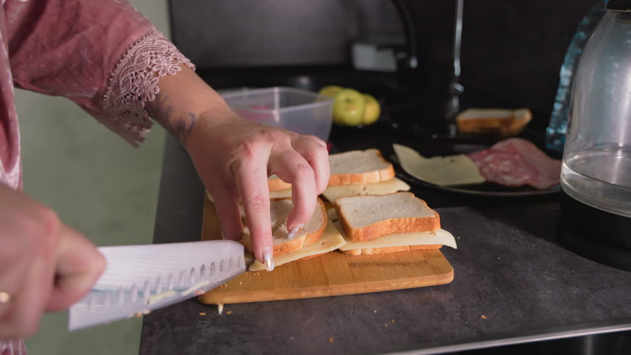 Caregiver in pink robe cuts sandwiches on kitchen counter using sharp knife, preparing breakfast with cheese, bread, and deli meat while surrounded by apples, containers, and electric kettle in cozy home setting