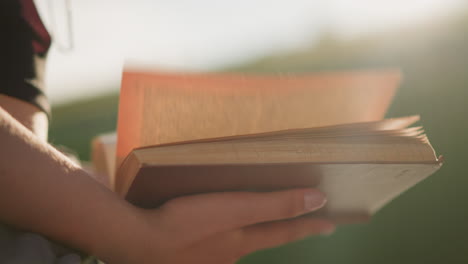 close-up of an individual holding and flipping through book pages to the right, sunlight softly reflects on the person and book, with a blurred background