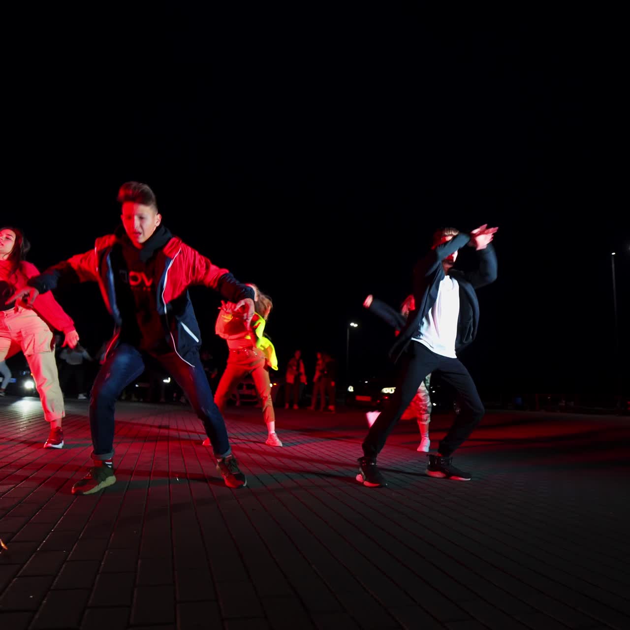 Two boys and three girls dancing hip hop for audience outdoors. Dance group performing in the lights of cars at backdrop. Low angle view