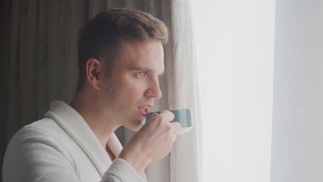 Man-Wearing-Bathrobe-On-Hotel-Or-Spa-Break-Standing-By-Open-Curtains-With-Hot-Drink