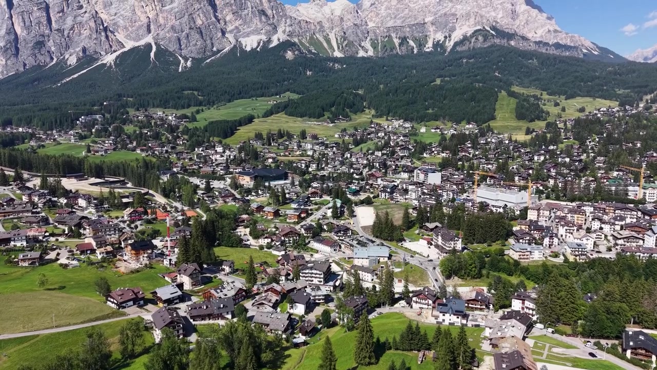 Drohnenansicht über Cortina d'Ampezzo, die seine Wohnviertel, grüne Wiesen, bewaldete Hänge, Straßen und die dramatischen Dolomiten im Hintergrund zeigt.