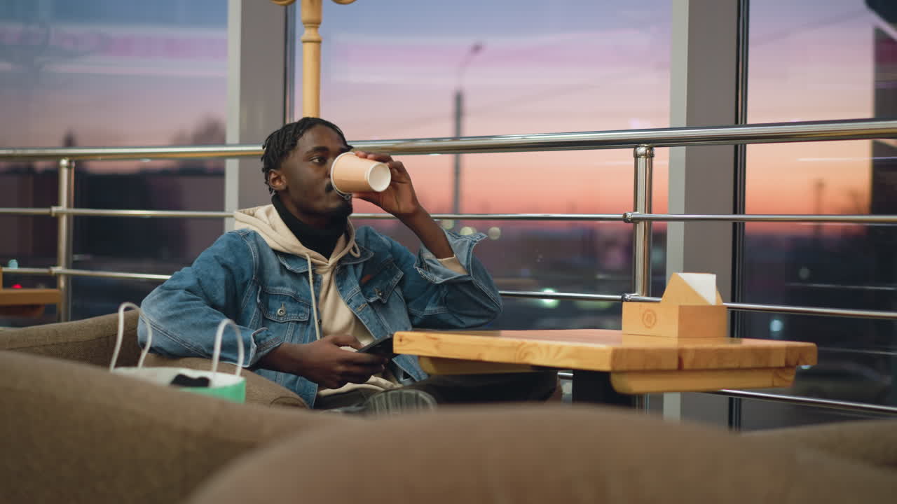Young man dressed in layered hoodie and denim jacket sits alone in cozy lounge during evening, calmly checking smartphone while reaching for coffee cup on wooden table, soft sunset sky glowing