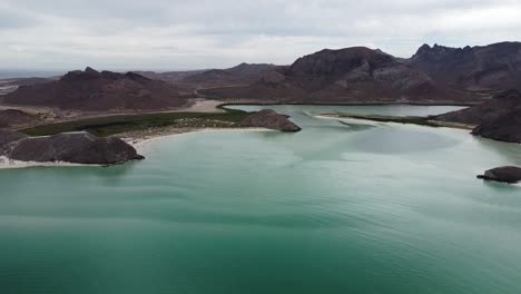 Playa-balandra-with-turquoise-waters-and-mountain-backdrop,-baja-california,-mexico,-cloudy-sky,-aerial-view