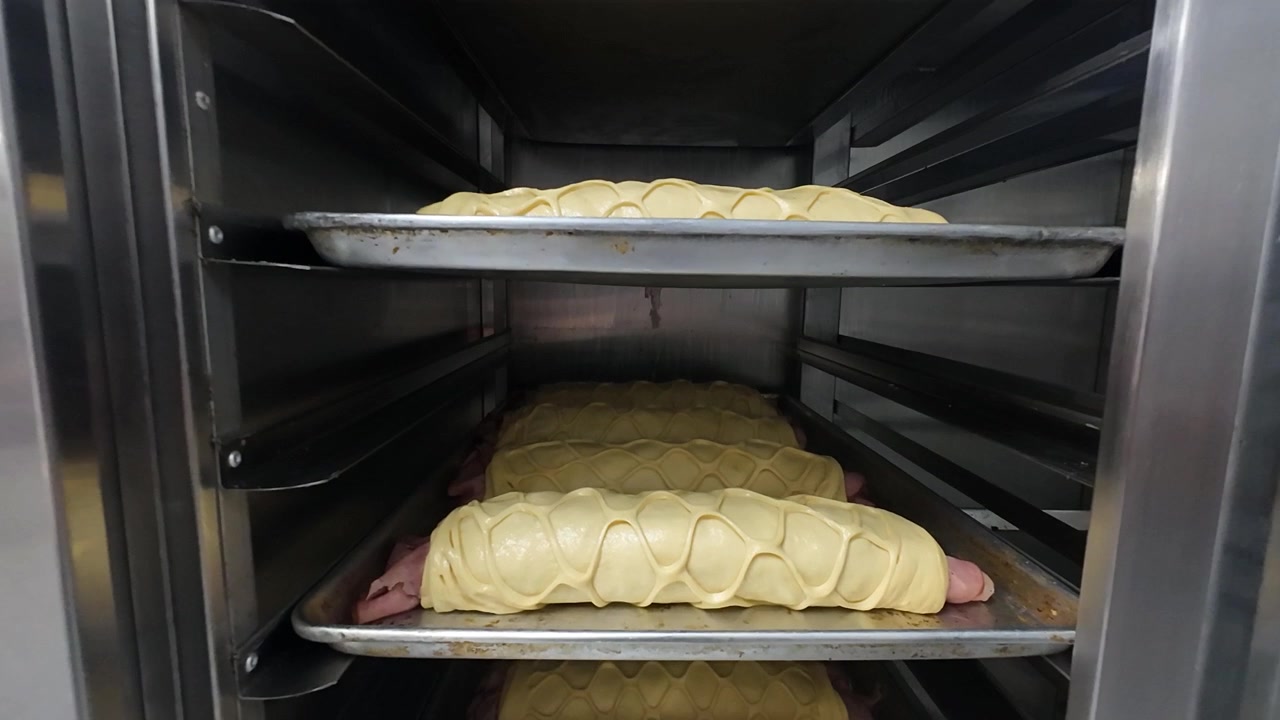Panorering nedover mot flere brett med tradisjonelt juleskinkebrød (Pan de Jamon) inne i en profesjonell industriovn. Deigen har klassisk kryssmønster, klar til steking i et bakeri i Caracas