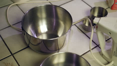 a kitchen counter covered in metal mixing bowls, measuring cups and cooking utensils as a chef prepares to bake a chocolate cake