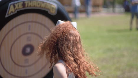 Cute-young-girl-with-red-hair-throwing-toy-axe-at-a-target-outdoors-in-a-park-midday