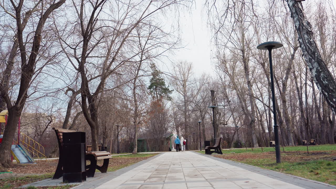 Lower view of people walking along tiled pathway surrounded by tall trees with dry leaves and empty benches in calm urban park, capturing quiet atmosphere of winter or late autumn