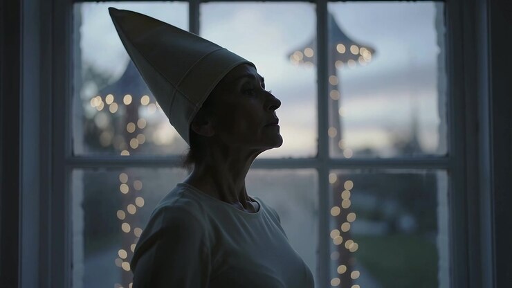 Sollevando la testa, una donna con un cappello conico osserva le luci attraverso una finestra a vetri mentre scende il crepuscolo