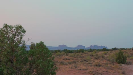 Amazing-mountain-rock-formations-sit-on-horizon-behind-shrubs---trees-Moab-desert,-Utah-dolly-shot