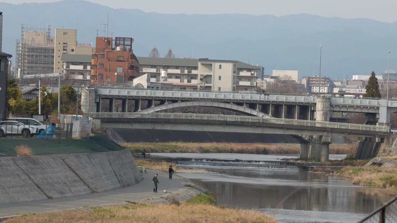 Inquadratura ampia statica di un ponte di cemento su un fiume con edifici urbani e montagne.