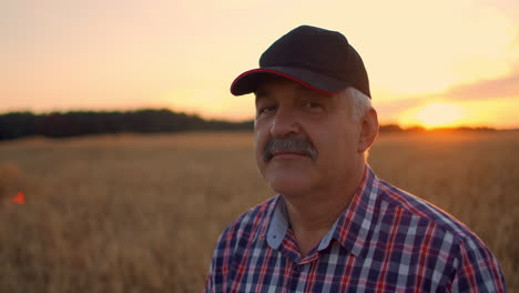 portrait of a happy senior adult farmer in a cap in a field of grain looking at the sunset. wheat field of cereals at sunset. slow motion