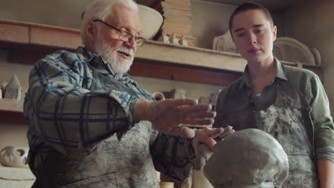 elderly sculptor teaching young apprentice in pottery studio