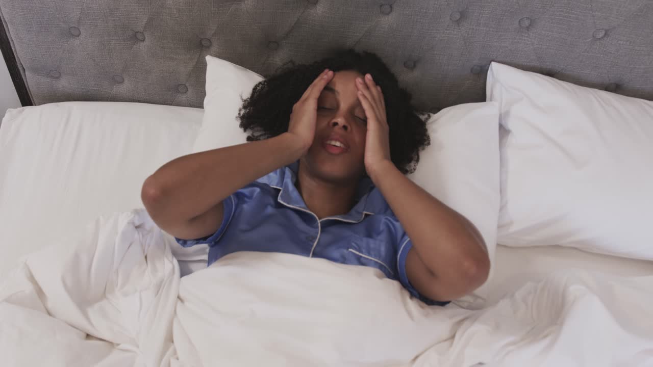 African American woman waking from sleep rubbing eyes massaging temples in bed wearing pajamas