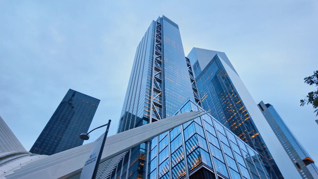Lavvinkelbillede af 3 World Trade Center og nabotårne, Lower Manhattan, New York. Opadvendt perspektiv viser glasfacader med diagonale stålafstivninger, Oculus-finner og træløv i skumringen