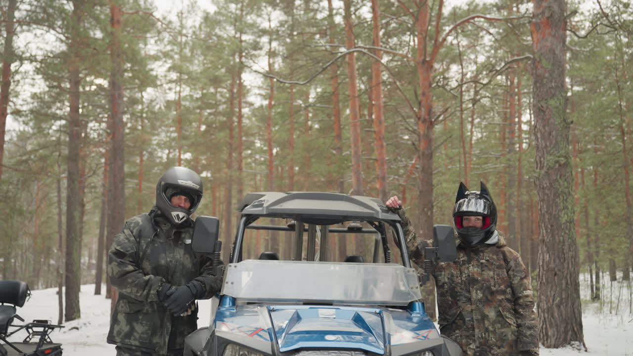 Due piloti accanto a un quad blu in una pineta innevata durante il briefing pre-corsa, indossano caschi e abbigliamento invernale, controllando il veicolo e discutendo il percorso e la sicurezza, creando un'atmosfera da spedizione fuoristrada avventurosa.