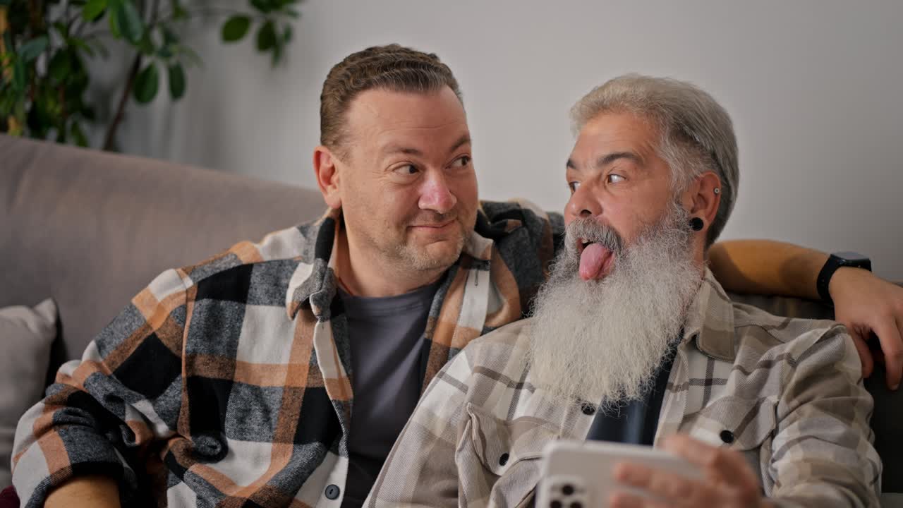 A happy brunette man with stubble in a checkered shirt together with his elderly boyfriend a man with gray hair and a lush beard communicate via video call and make faces while sitting on the sofa in a modern apartment
