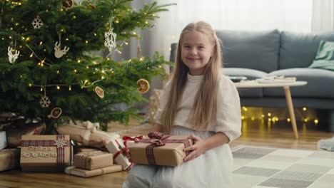 Caucasian-girl-holding-Christmas-gift-while-sitting-next-to-Christmas-tree.