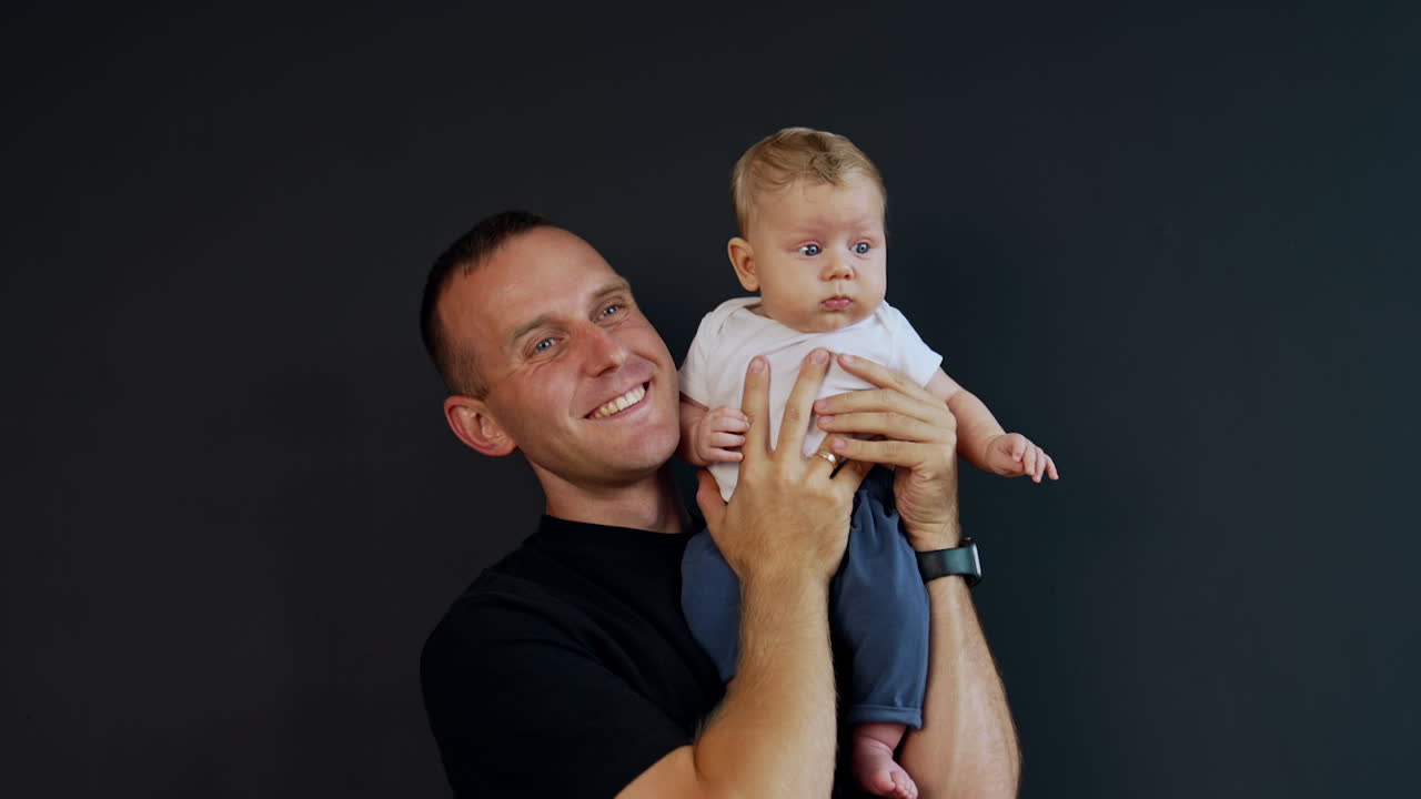 Happy male wearing black t-shirt holding his infant son on the shoulder. Little baby looking around and father kisses him on the cheek.