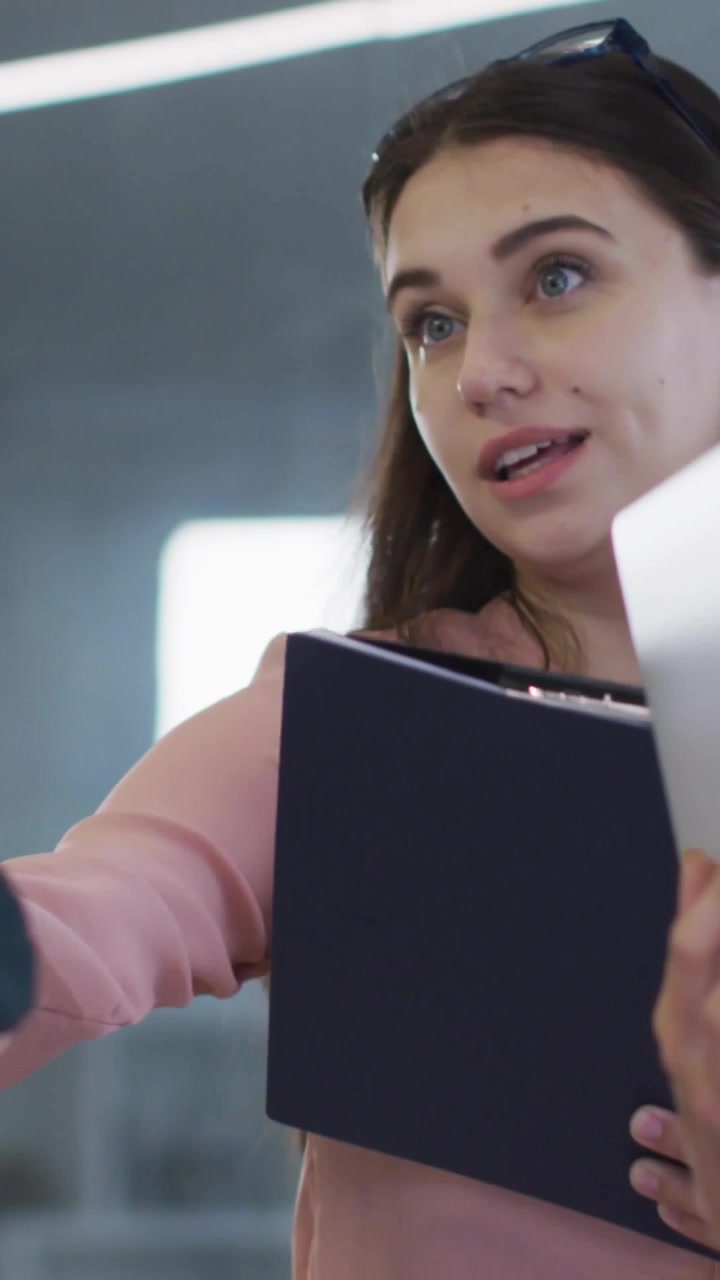 Video verticale: Donna con top rosa che prende documenti di lavoro da un collega che si protende, mentre sovrapposizioni di lavoro oscurano la scena.