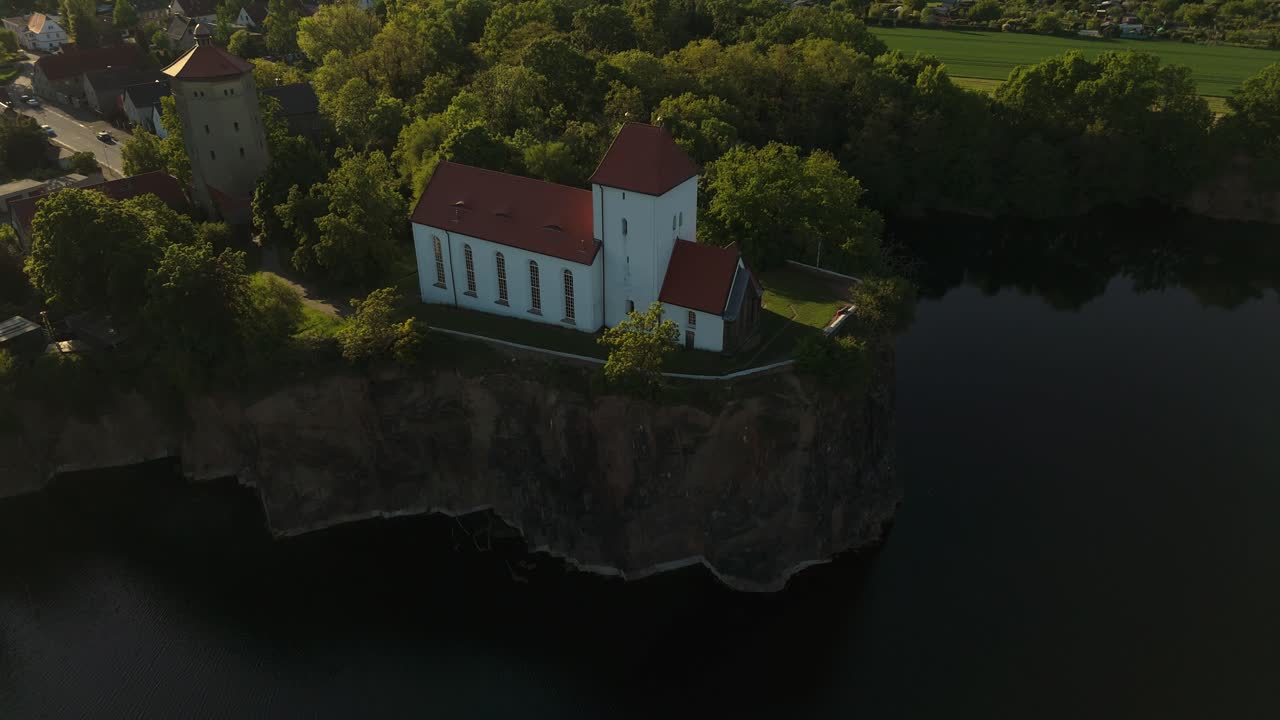 Cinematic drone shot of the Wehrkirche Beucha near Leipzig at sunset. The historic church sits dramatically on a cliff above the flooded quarry, surrounded by forest and glowing evening light.