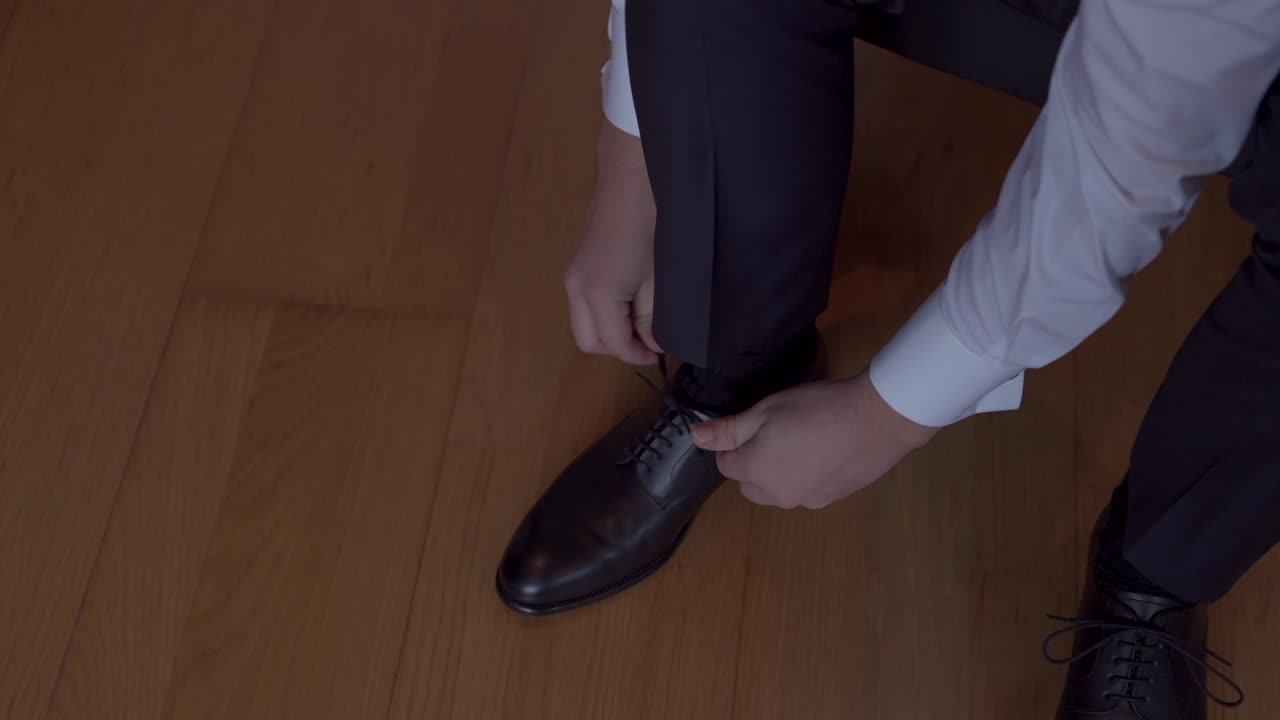 Close up of groom tying black dress shoes over polished wooden floor
