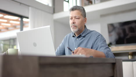 Fokussierter-älterer-Mann-Mit-Gemischter-Abstammung,-Der-Am-Tisch-Sitzt-Und-Auf-Den-Laptop-Schaut,-Von-Zu-Hause-Aus-Arbeitet,-Zeitlupe