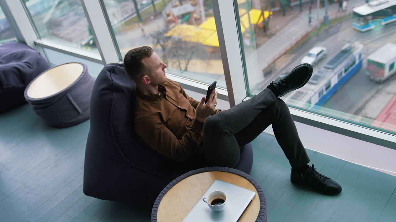 Businessman relaxing on view from office window. Guy with a phone looking at city through window on comfortable ottoman. Work life balance concept. Top view.