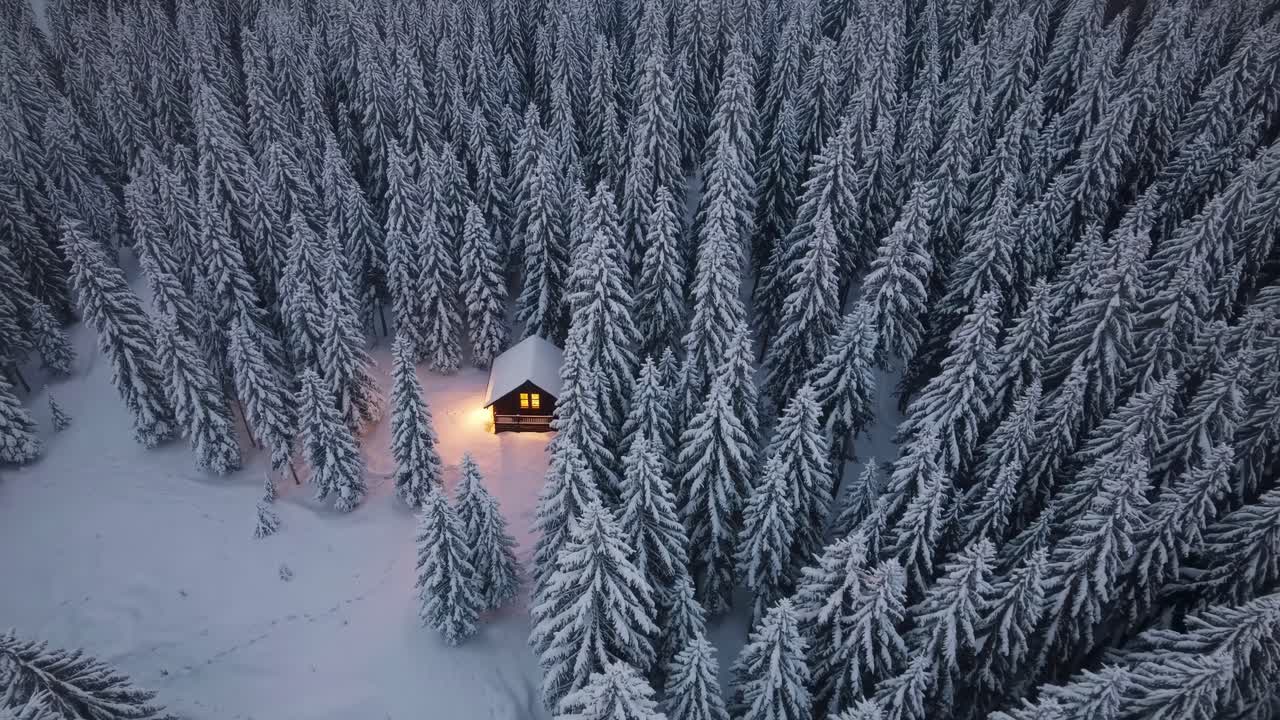 Aerial view of a snow-covered forest with a cozy cabin illuminated by warm light, creating a serene