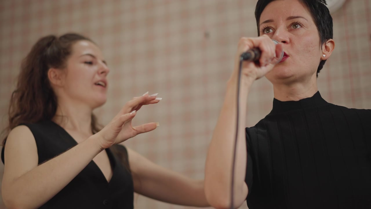 Supportive duet coaching with white women, older singer receives hands on guidance from young assistant, microphone handover and shoulder touch for posture, classroom rehearsal, warm rapport, harmony