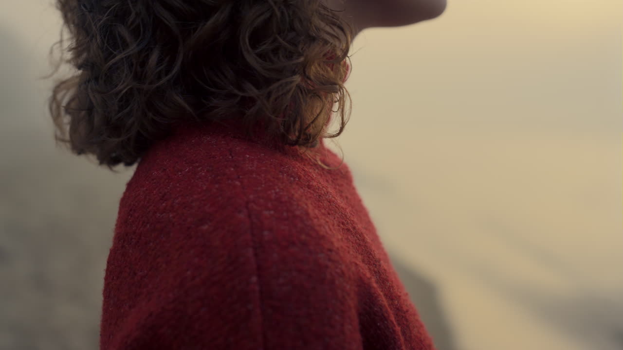 Kvinde står på havstrand ved solopgang. Nærbillede af stilfuld pige iført rød sweater.