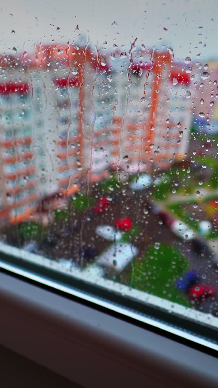 View through the window on raining weather. Drops of rain on a window pane on the city buildings background. Rain droplets flowing on a glass. Vertical video