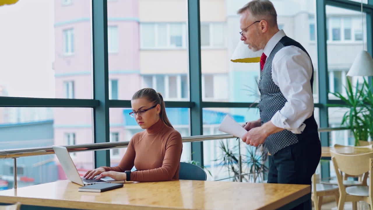 Young woman working on a computer near the window. Senior businessman coming to his secretary sitting at the desk and showing her some papers. Slow motion.
